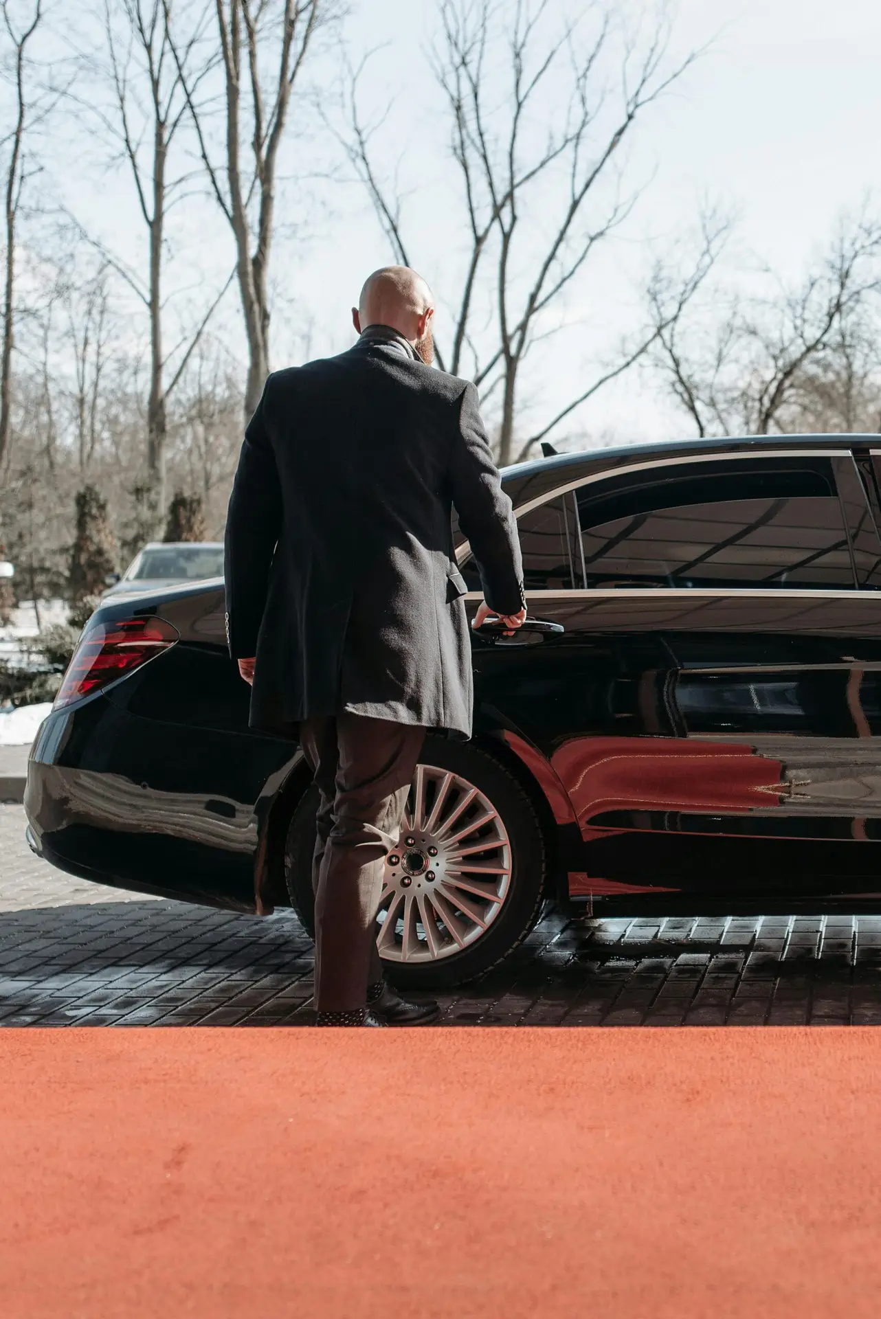 Bald man in a black suit approaches a luxury black car on a sunny day