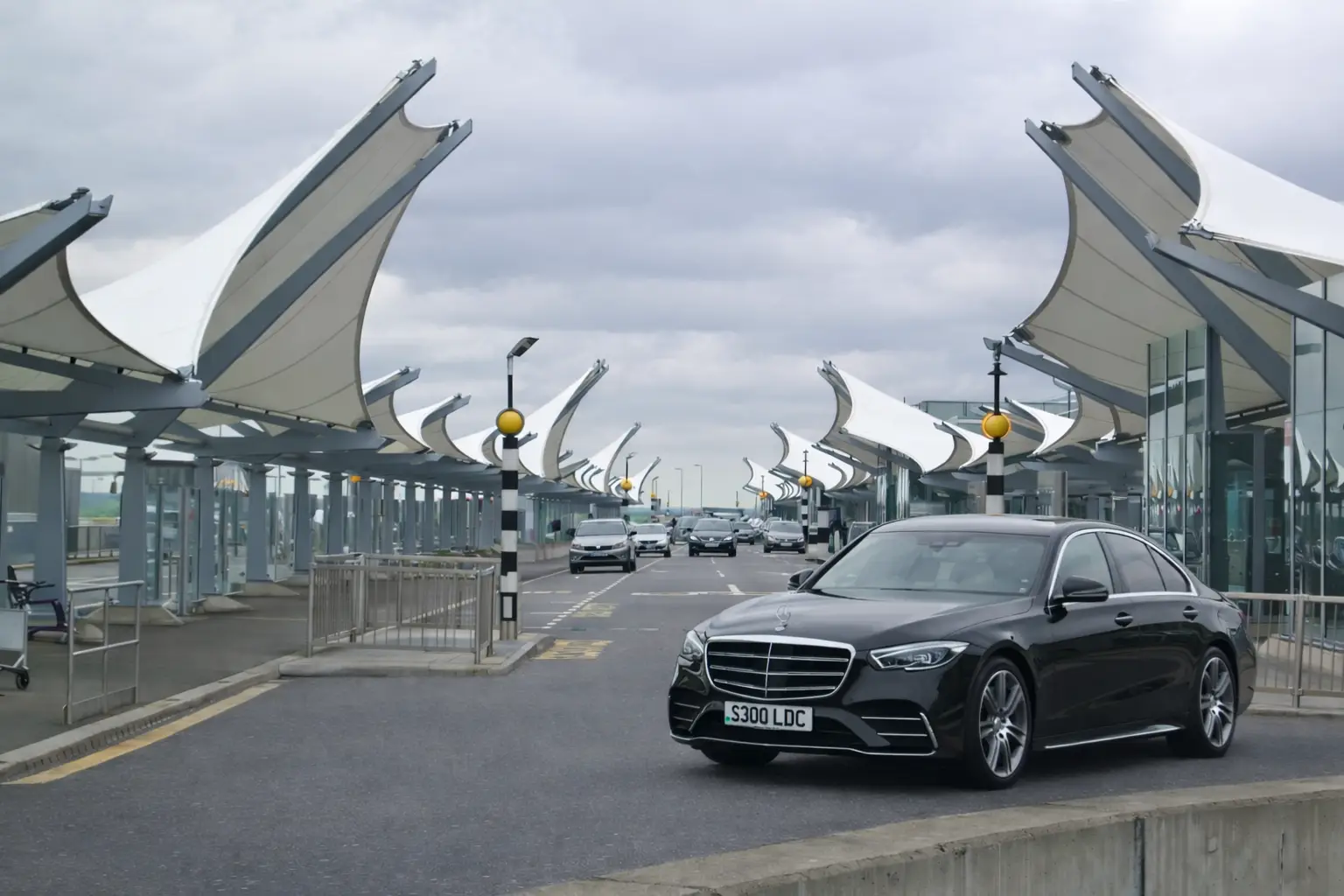 Black Mercedes S Class chauffeur car at airport drop off with modern terminal architecture.
